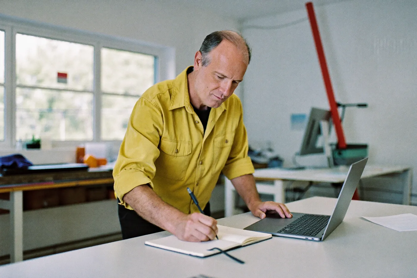 A man writing in a paper notebook beside a closed laptop in a workshop office
